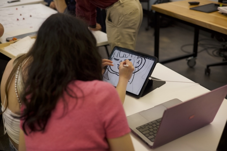 Photo taken behind two Tisch Summer High School Game design students. One student draws on a tablet while the other one watches over her shoulder.